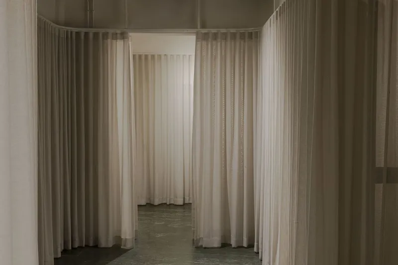 Narrow passageway formed by overlapping sheer curtain panels in neutral tones over polished concrete floor
