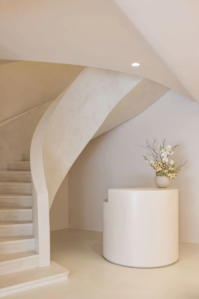 Curved staircase with pale stone treads rising beside a cylindrical plinth holding a floral arrangement