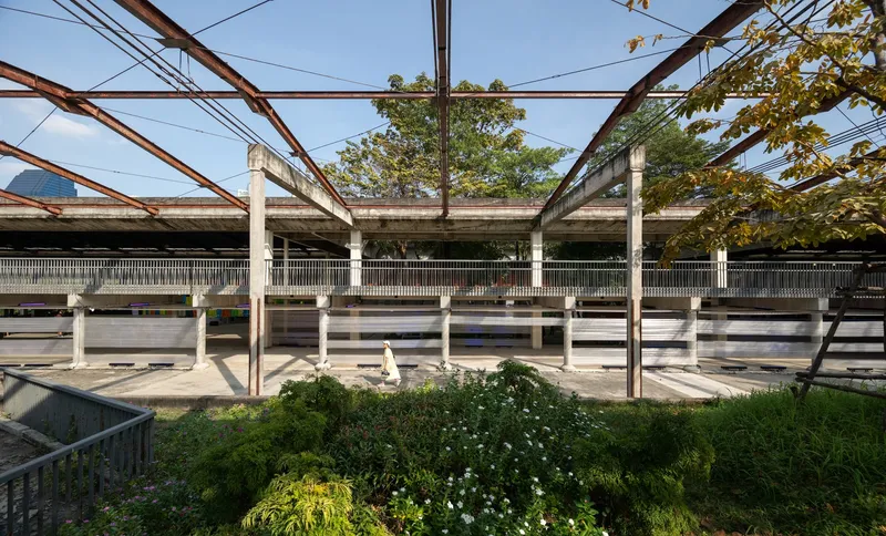 Covered outdoor platform with concrete columns and planted beds beneath a metal truss canopy