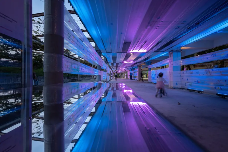 Covered corridor with reflective floor illuminated by purple and blue light strips at dusk