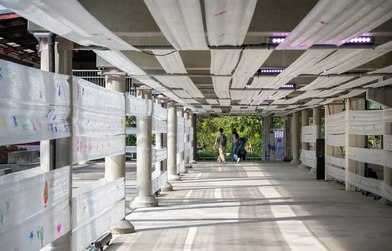 Covered walkway with radiating pleated ceiling panels, white fabric screens, and cylindrical columns as pedestrians pass through