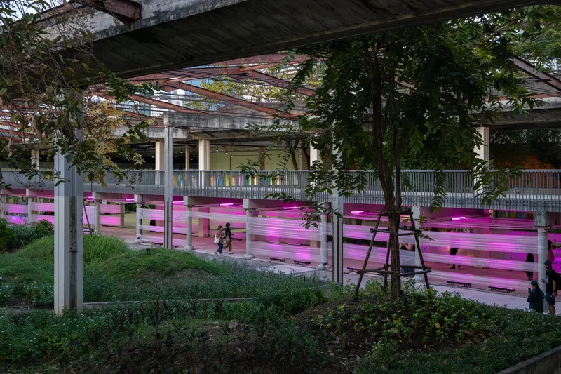 Magenta-lit horizontal screening panels with pedestrians passing beneath steel trusses and mature trees