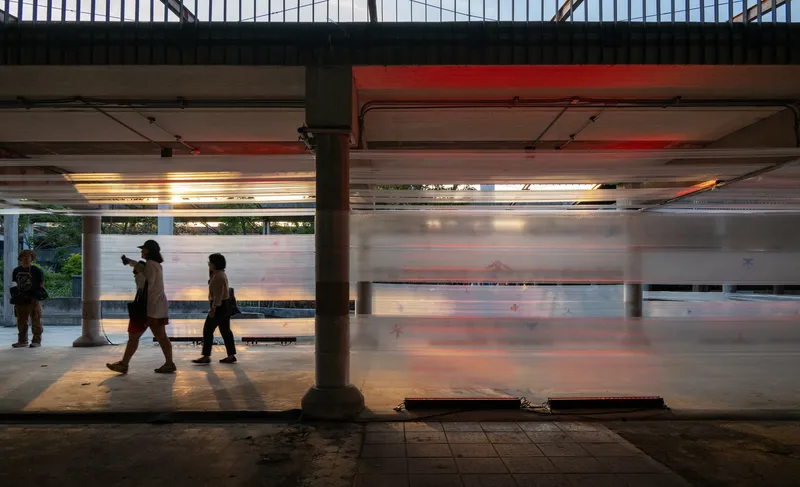 Platform edge with concrete columns and pedestrians walking past translucent backlit wall panels at dusk