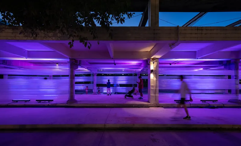 Covered pedestrian walkway with purple LED lighting along concrete columns and a person in motion at dusk