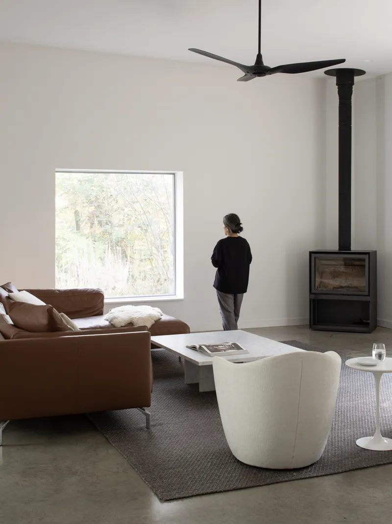 Living room with freestanding wood stove and person standing at a square window framing forest view