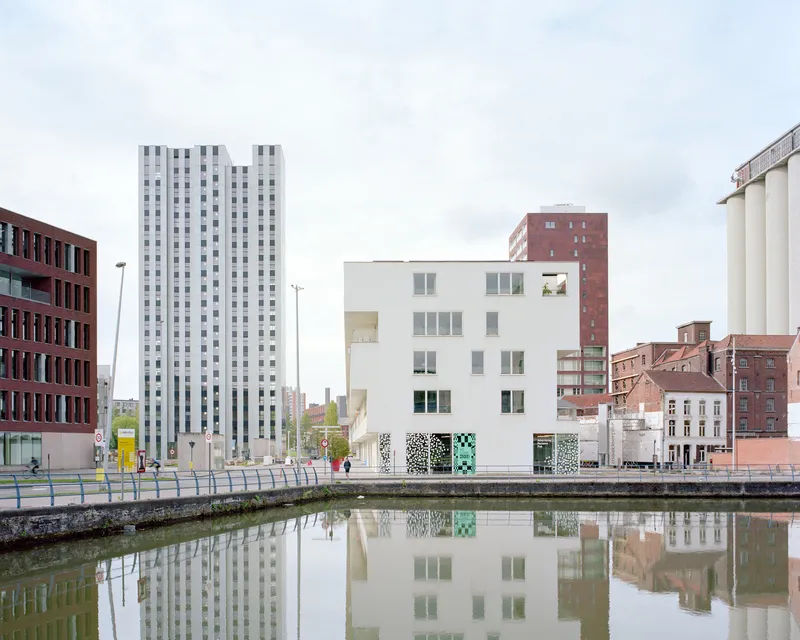 White cubic volume with patterned ground floor panels reflected in a calm canal under overcast skies