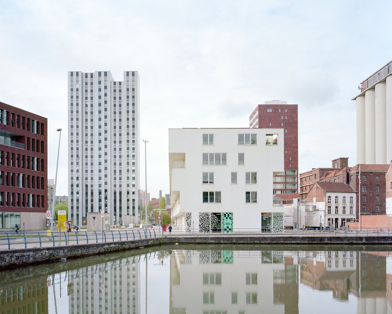 White cubic volume with patterned ground floor panels reflected in a calm canal under overcast skies