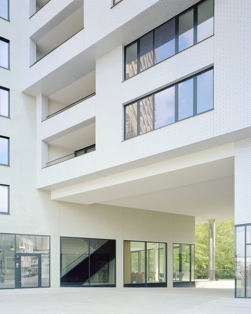 White brick facade with recessed balconies and a covered ground-level entry plaza opening to green trees