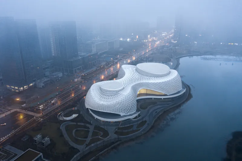 Three-lobed building with perforated white shell rising from the waterfront in heavy fog