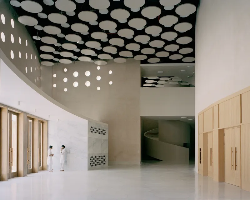 Interior lobby with perforated ceiling panels, timber doors, and two visitors standing near the elevator bank