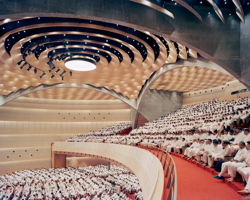 Auditorium interior with spiraling timber ceiling and tiered seating filled with attendees in white garments