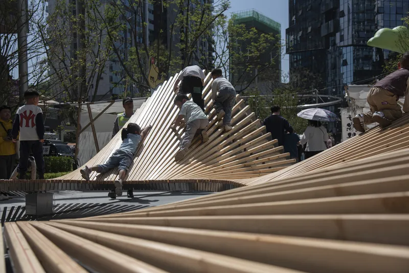 Undulating timber slat pavilion with children climbing on it in a sunny urban plaza