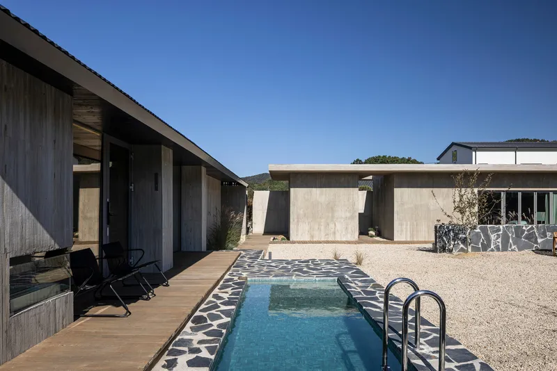 Lap pool with basalt stone edging between timber deck and gravel courtyard under clear sky