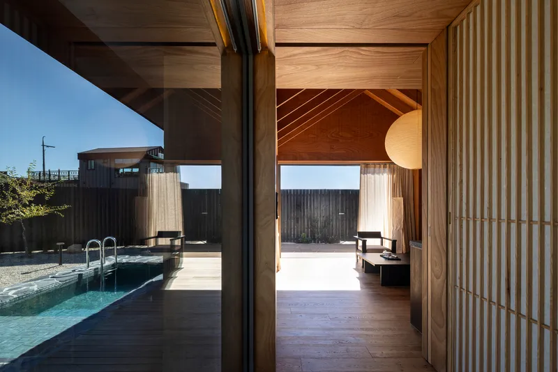 Interior view through timber screens toward a pool deck and covered outdoor space under clear sky