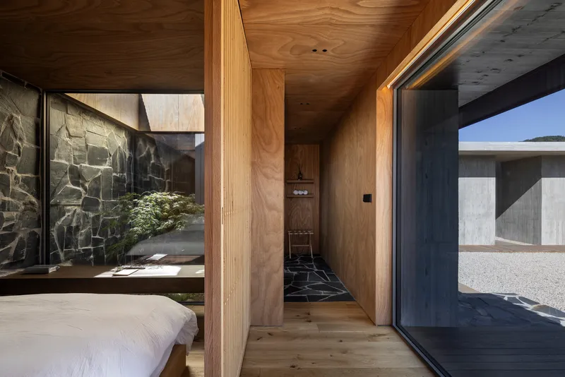 Timber-lined bedroom opening onto a courtyard with dark stone wall and planted greenery in morning light