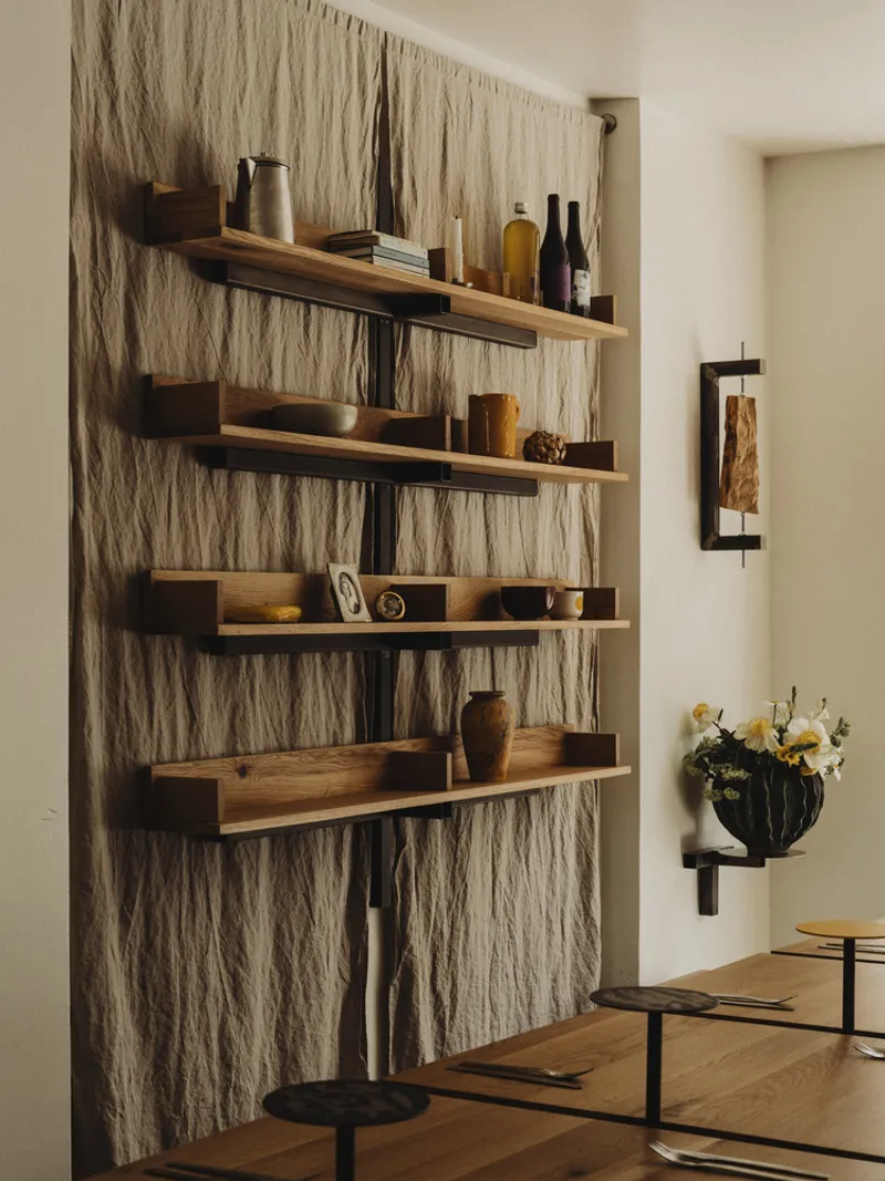 Wall-mounted timber shelves displaying ceramics against textured fiber panel with a vase of white flowers below