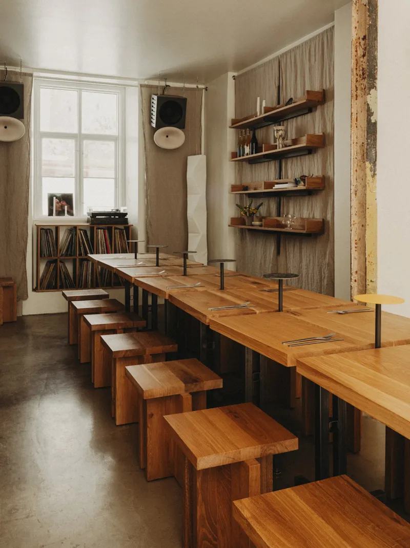 Communal timber dining counter with block stools and floating shelves near tall windows