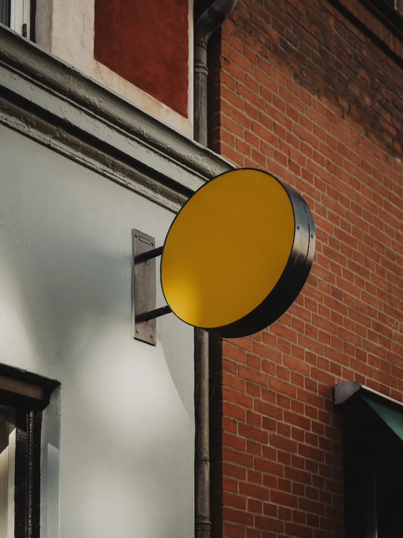 Circular yellow signage disc mounted on a white facade corner against red brick in daylight
