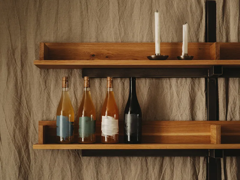 Timber shelving against a textured fabric wall displaying wine bottles with leather sleeves and white candles