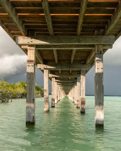 This architectural image showcases a wooden pier extending over calm, turquoise waters. The pier's s