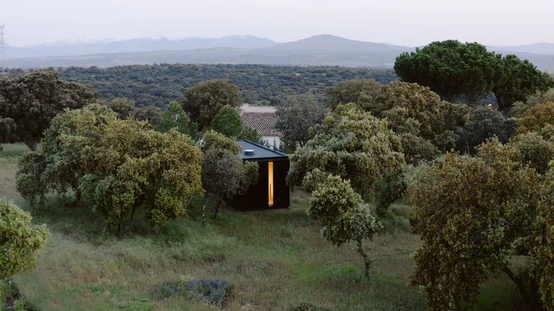 Dark timber pavilion nestled among olive trees with illuminated doorway at dusk beneath distant mountains