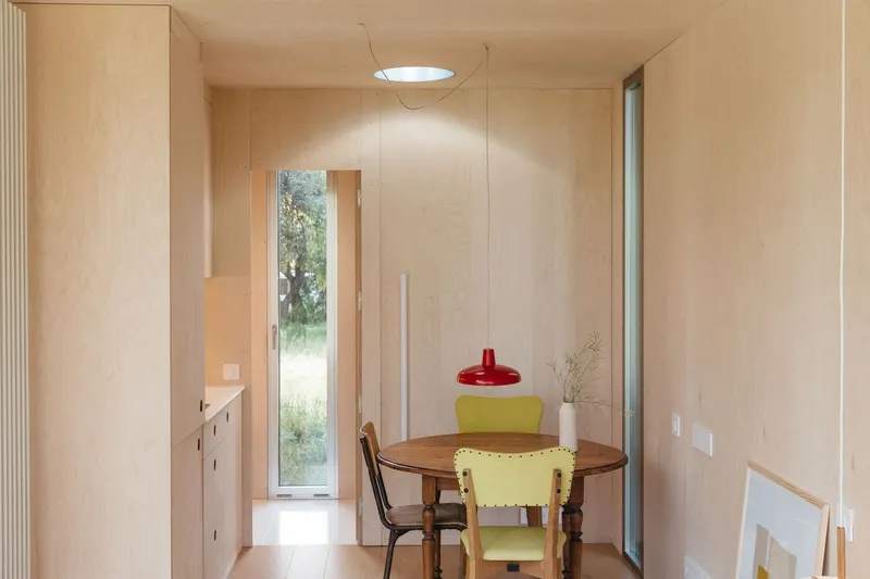 Light plywood-lined dining alcove with round table and narrow window opening to landscape beyond