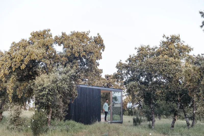 Charred timber-clad volume with glazed corner and figure in doorway among scattered oak trees