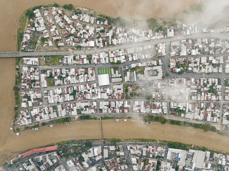 Aerial view of dense residential blocks along a winding river with floating structures at the water's edge