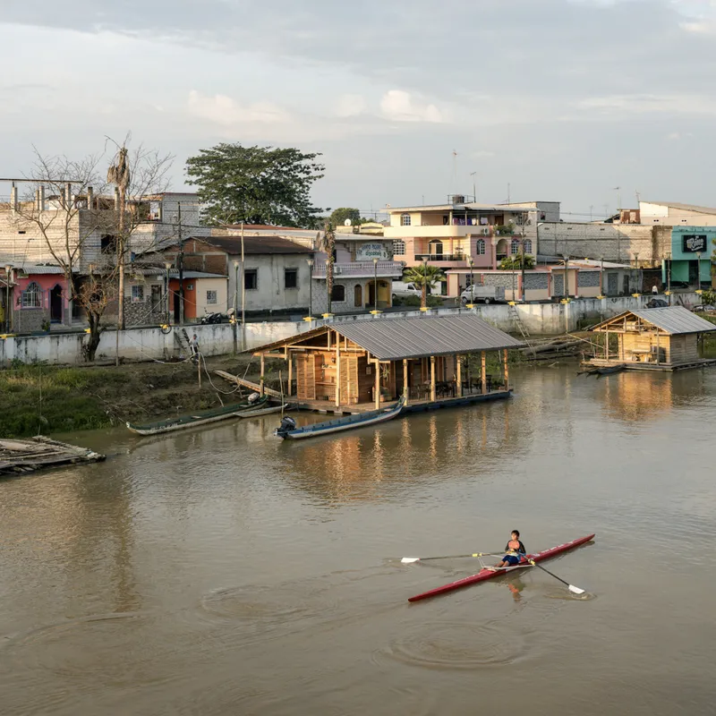 Two floating timber pavilions moored along the riverbank at dusk with a rower passing in the foreground
