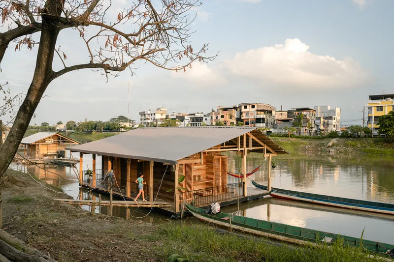 Floating structure with timber louvers and metal roof moored at grassy riverbank under afternoon clouds
