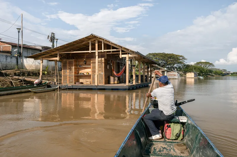 Timber-framed floating house with slatted wood walls viewed from a canoe on a muddy river