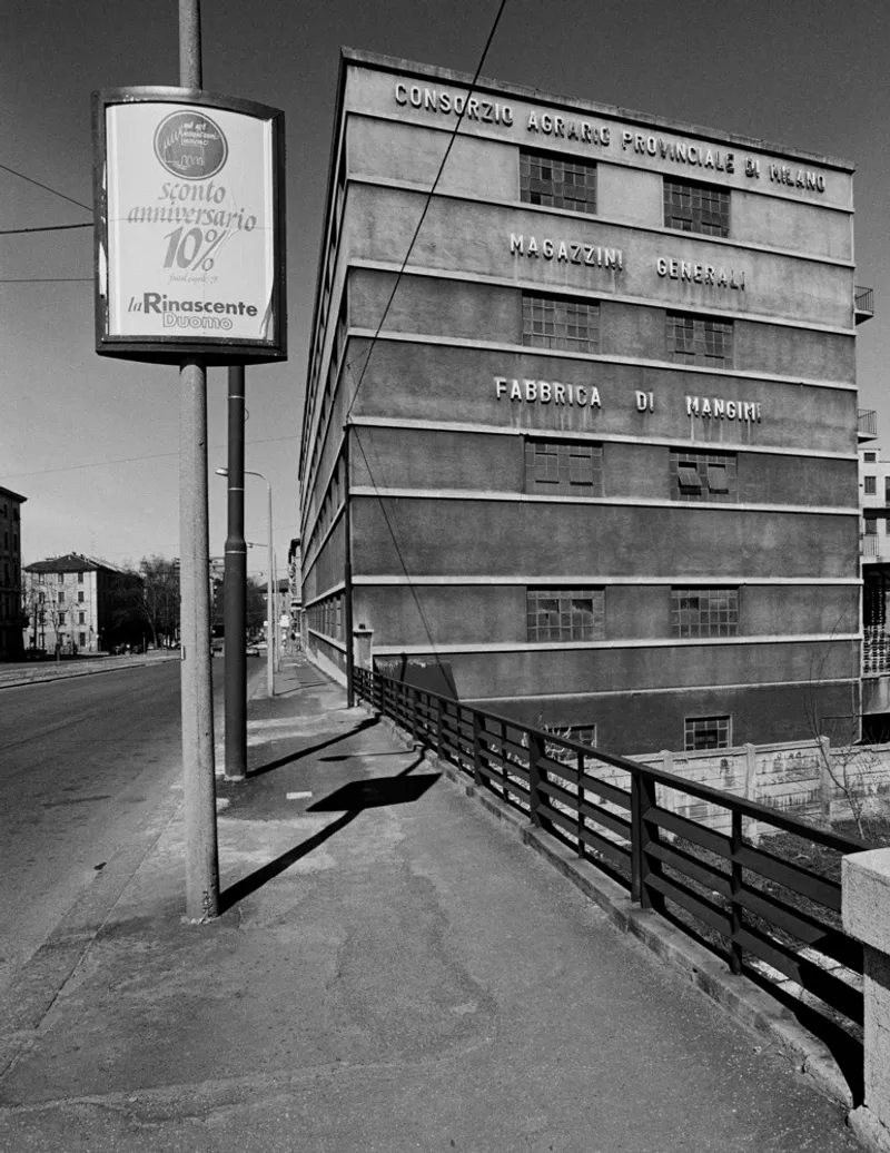 Horizontal banded facade with lettering alongside a sidewalk with advertising sign and wooden fence