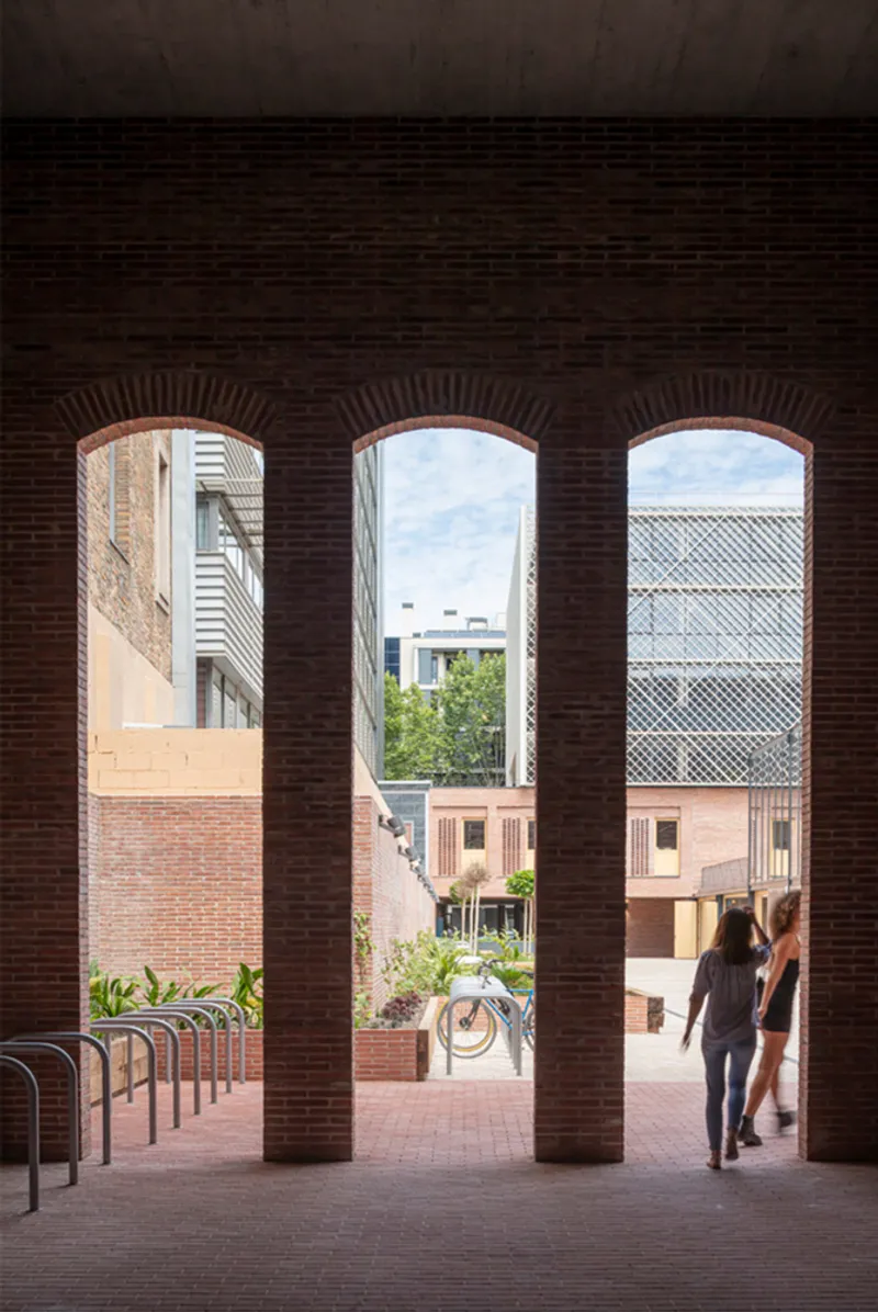 Three brick arches framing views of courtyard and surrounding buildings with two figures walking through