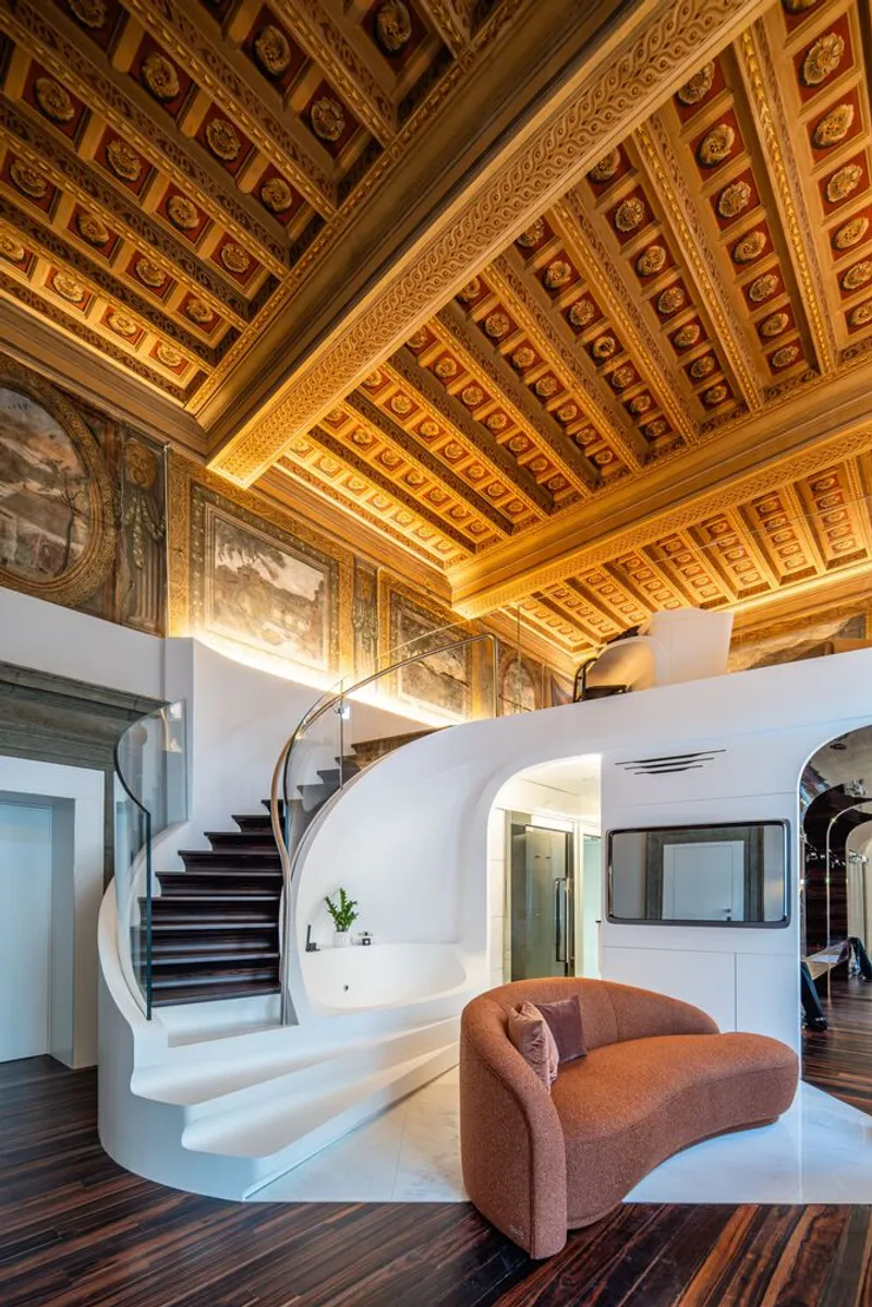 Curved white staircase rising beneath an ornate coffered ceiling with gilded details and historic frescoes