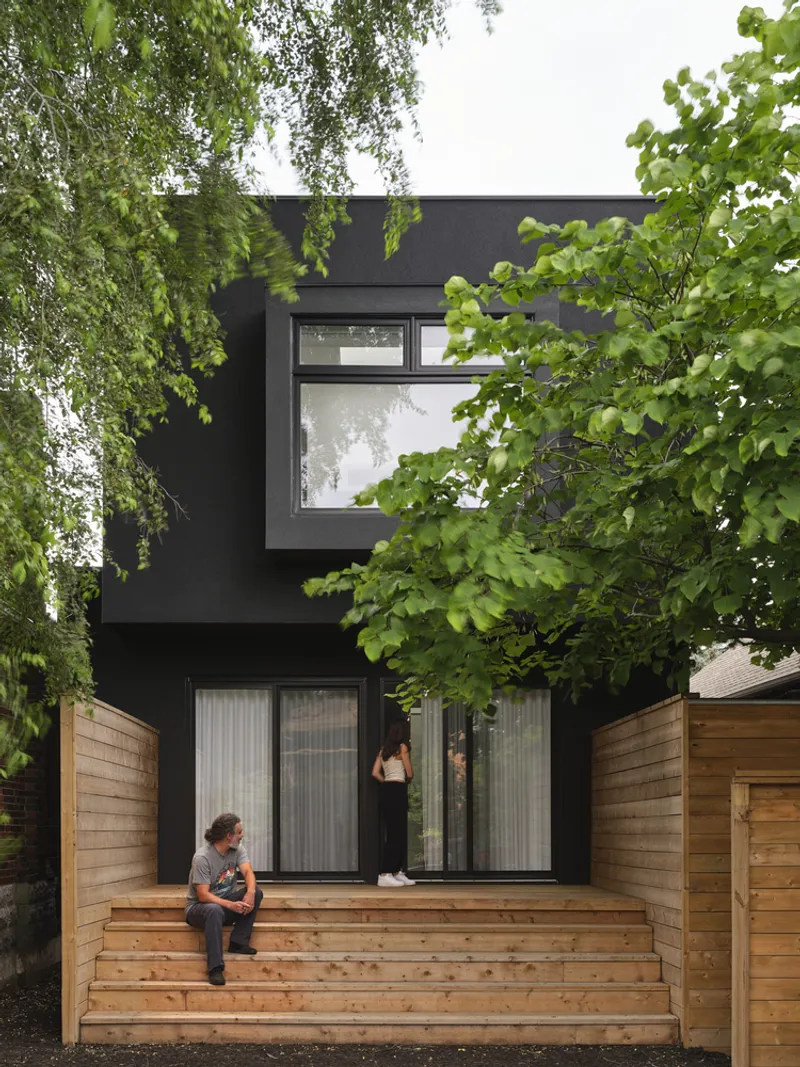 Black facade with timber stair and deck framed by green deciduous trees and wood fencing