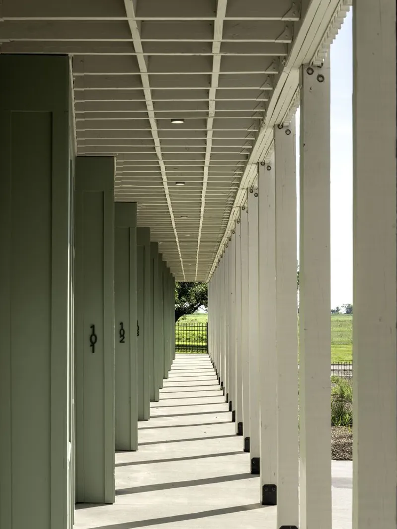 Covered walkway with white columns, slatted ceiling, and numbered green doors opening to landscape beyond