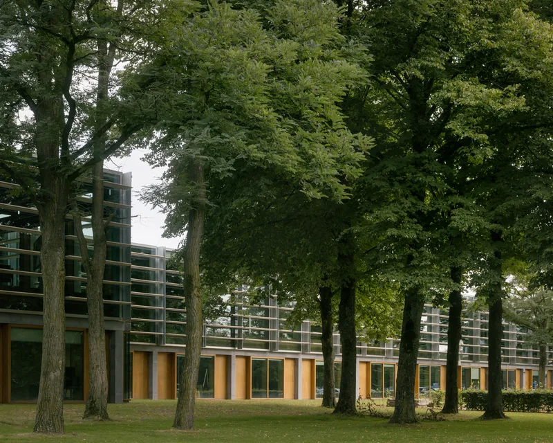 Long facade with horizontal louvers and timber glazed bays set within a park landscape