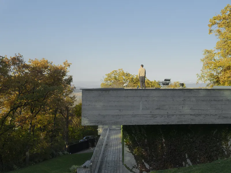 Cantilevered concrete volume on pilotis with figure on roof and autumn trees beyond