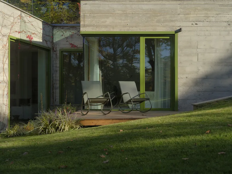 Board-formed concrete facade with chartreuse-trimmed glazing and two rocking chairs on a timber deck at dusk