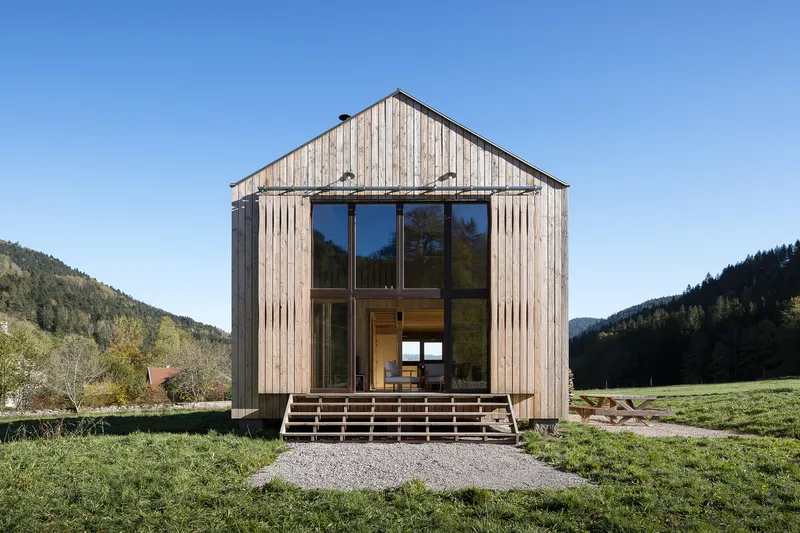 Vertical timber-clad facade with full-height glazing set in a meadow with forested hills behind