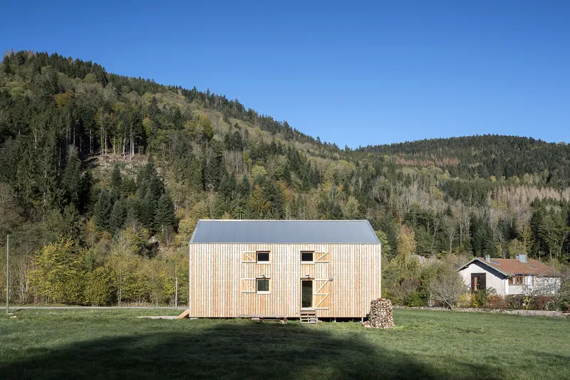 Side elevation showing vertical timber cladding and window openings with stacked firewood under clear autumn sky