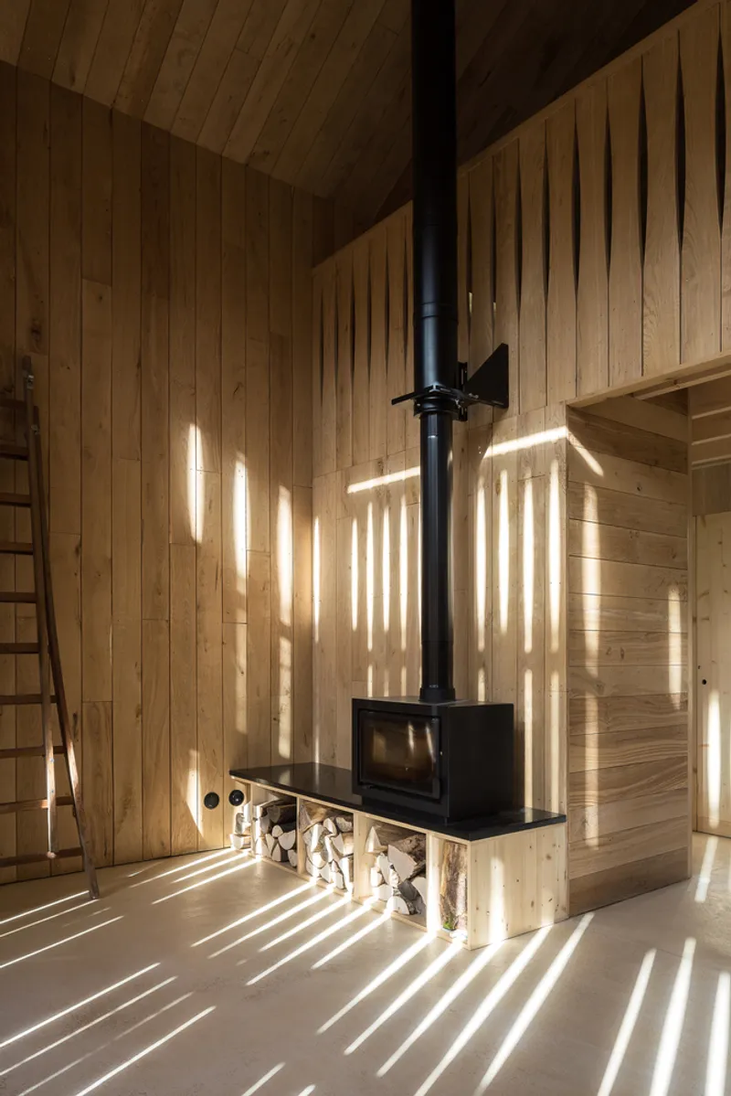 Wood stove with stacked firewood below casting afternoon light patterns across timber walls and concrete floor