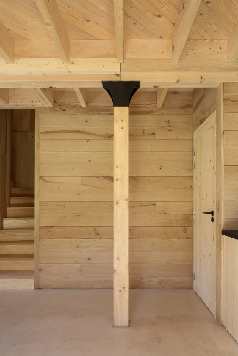 Timber column with black junction detail beneath exposed ceiling beams in hallway with staircase and doorway
