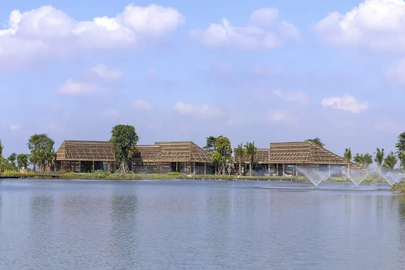 View across the pond showing three low pavilions with thatched roofs and fountain spray under summer clouds