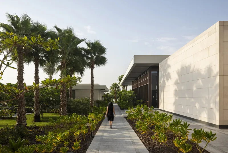 Garden pathway flanked by palm trees leading to a pale stone pavilion with vertical timber louvers