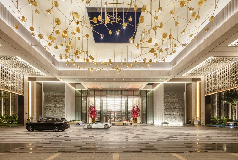 Porte-cochère with suspended brass pendant installation and cars beneath patterned stone ceiling at dusk