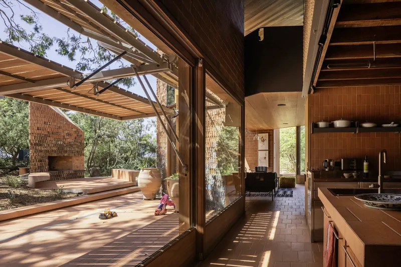 Kitchen with folding timber doors opening to a covered outdoor deck and brick fireplace under dappled light