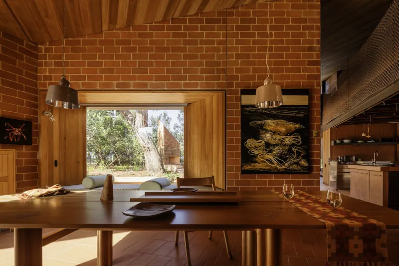 Dining area with timber table and window framing eucalyptus trees beyond red brick walls