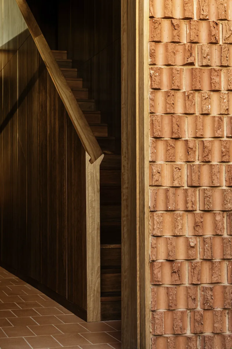 Timber staircase alongside terracotta brick wall in afternoon sunlight filtering through opening