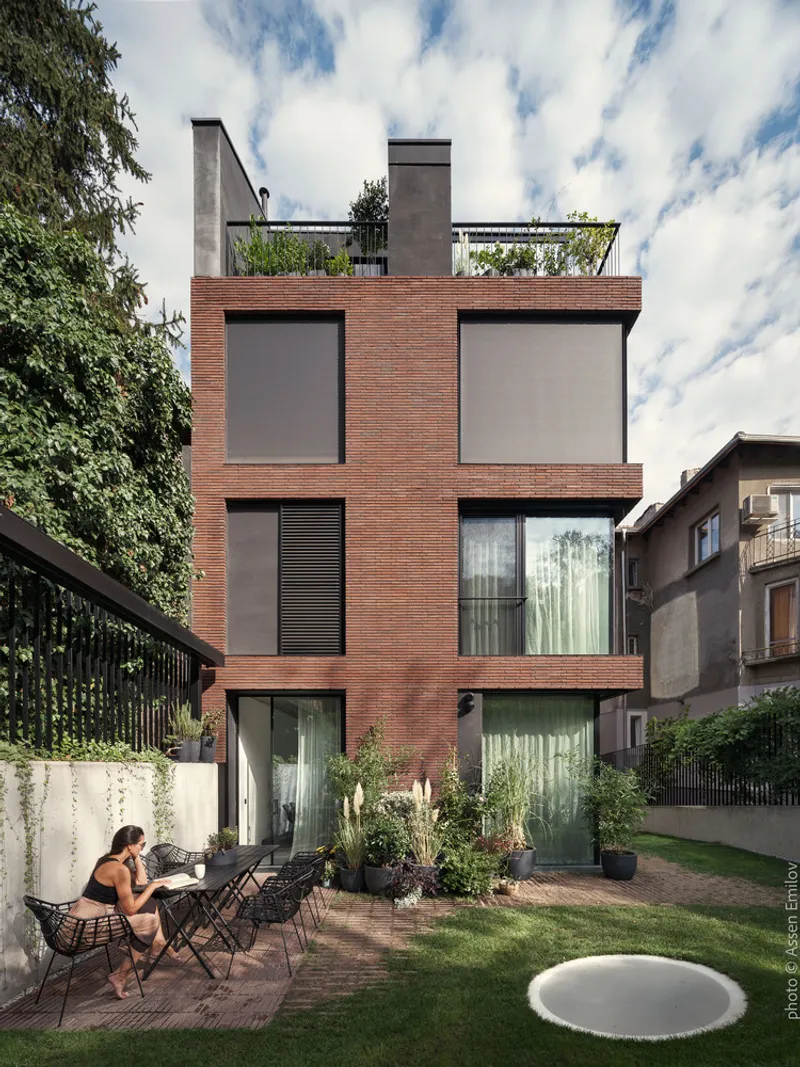Garden facade of the red brick volume with a woman seated on the timber deck surrounded by lawn and plantings